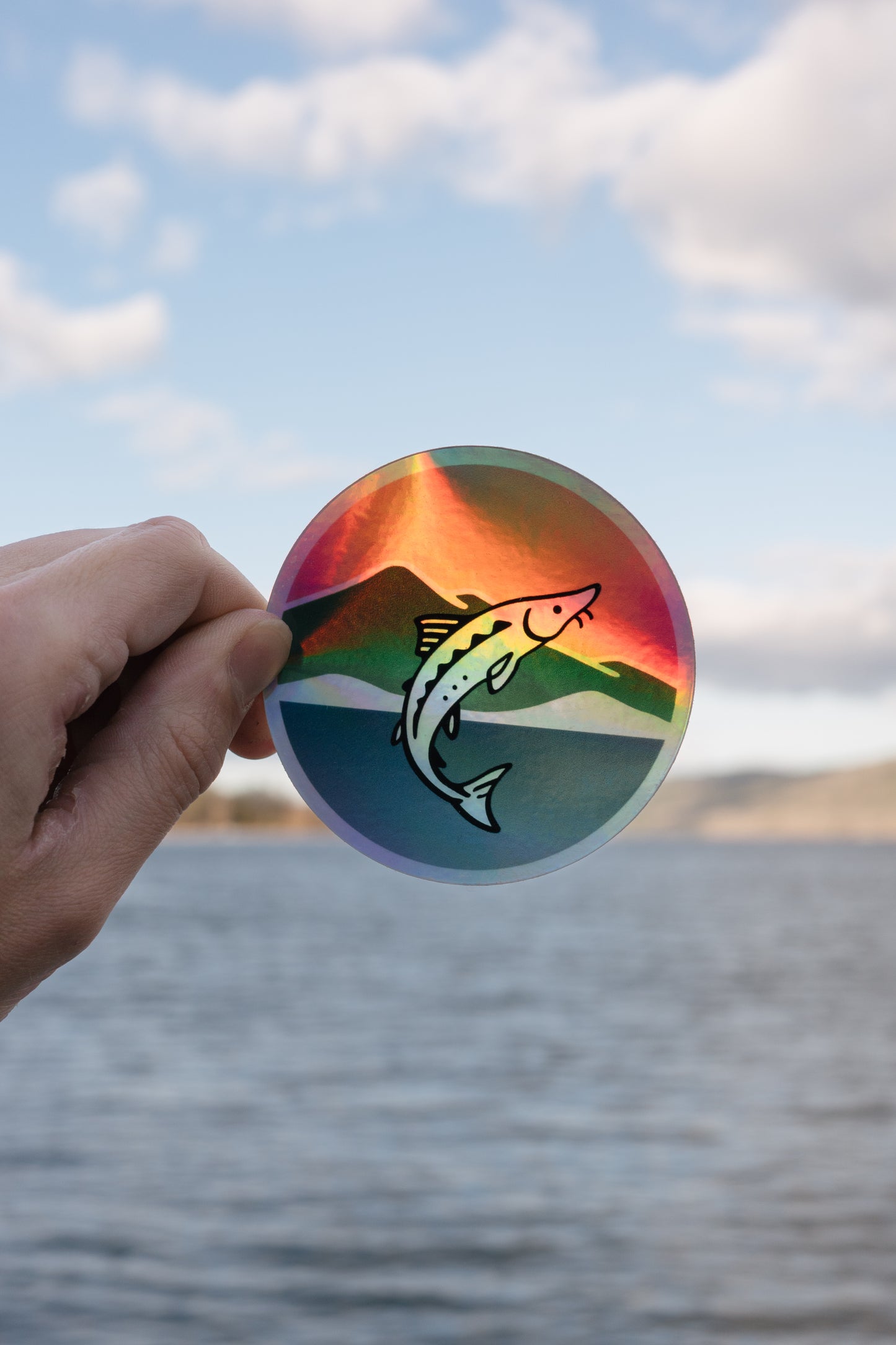 Hand holding a colorful holographic sticker with a sturgeon design against a water and sky background